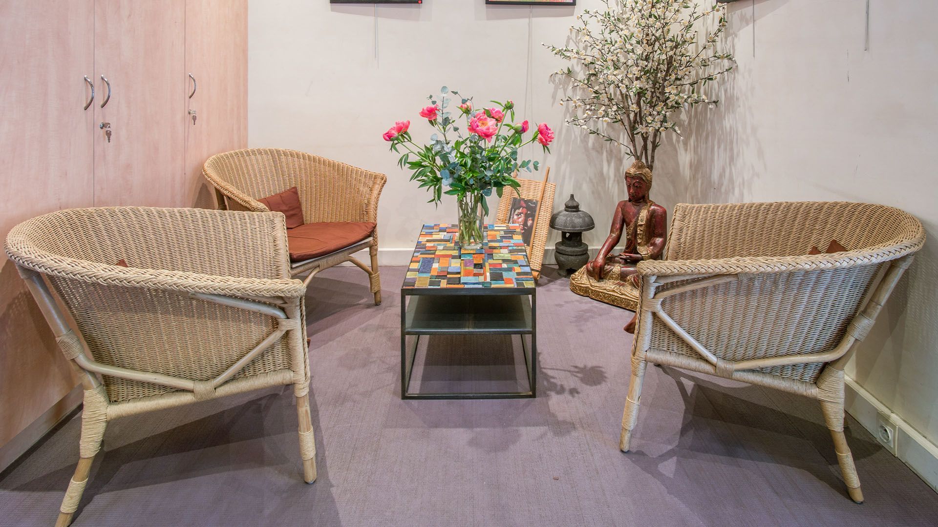Des chaises en osier disposées autour d'une petite table avec des fleurs, une statue de Bouddha et une armoire dans une pièce confortable.