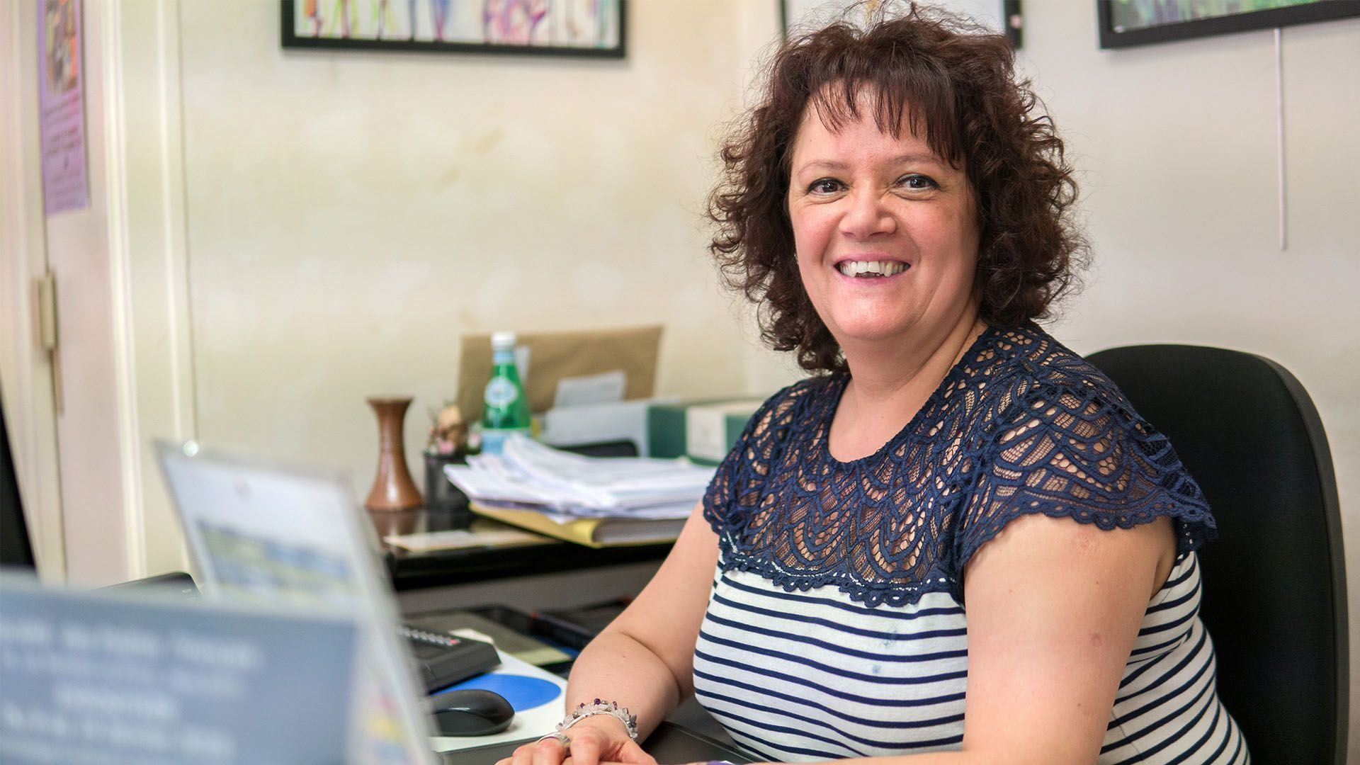 Une femme sourit à son bureau, dans un environnement de bureau. Elle porte une chemise à rayures.