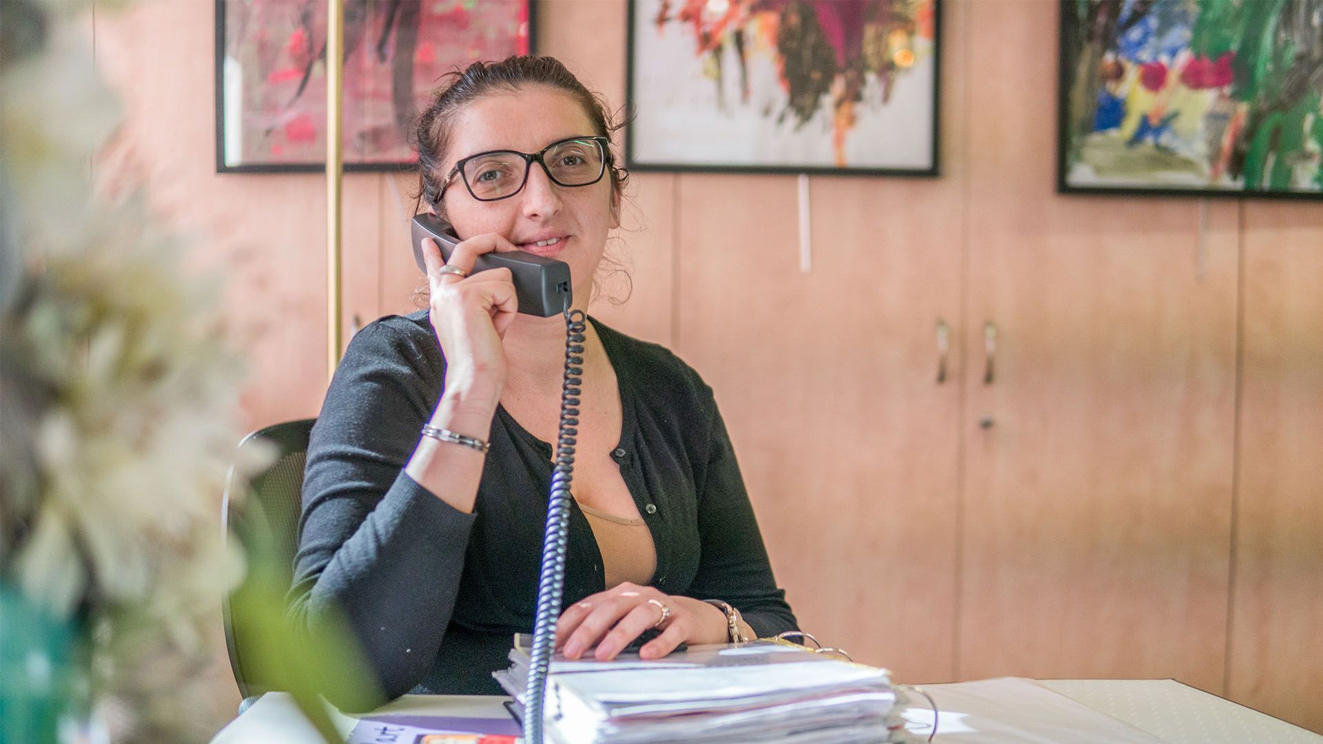 Une femme portant des lunettes parle au téléphone, assise à un bureau avec des papiers. Des œuvres d'art sont accrochées au mur.