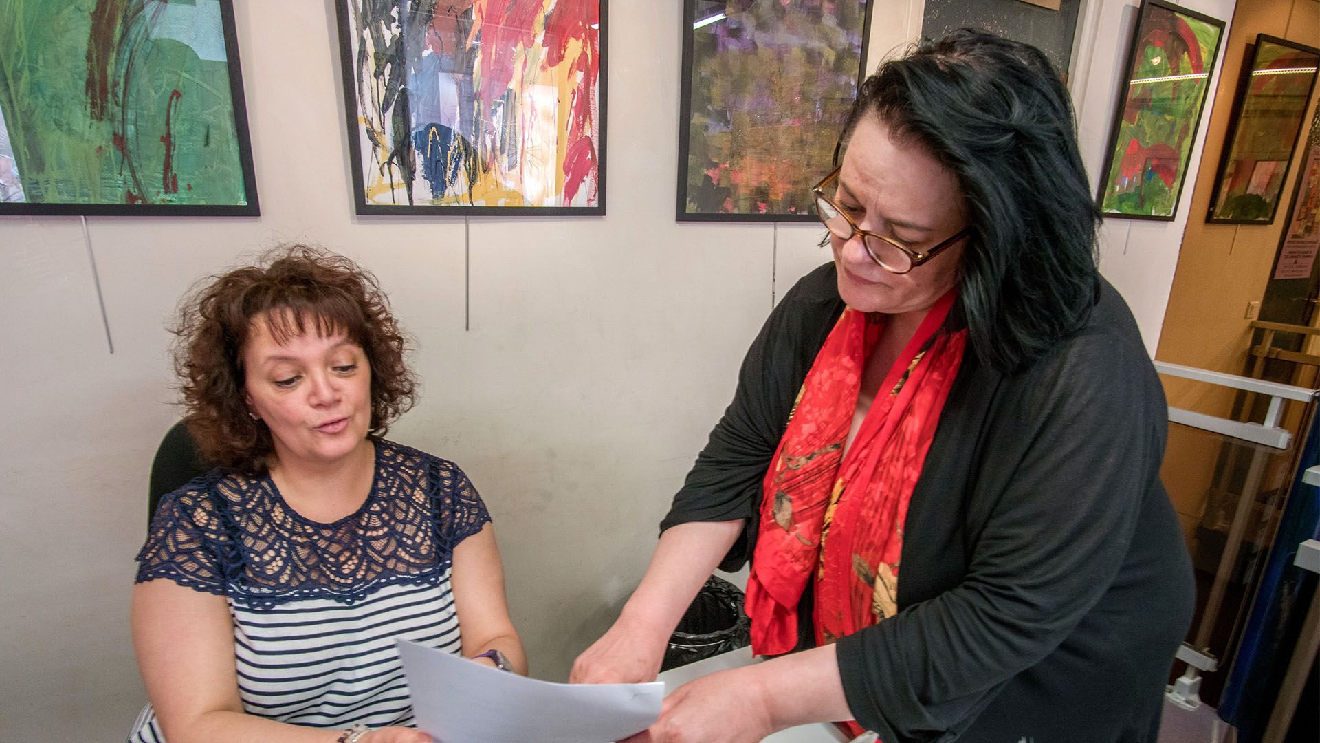 Deux femmes examinent des documents dans une galerie. Celle de droite désigne un document ; plusieurs tableaux sont accrochés au mur.