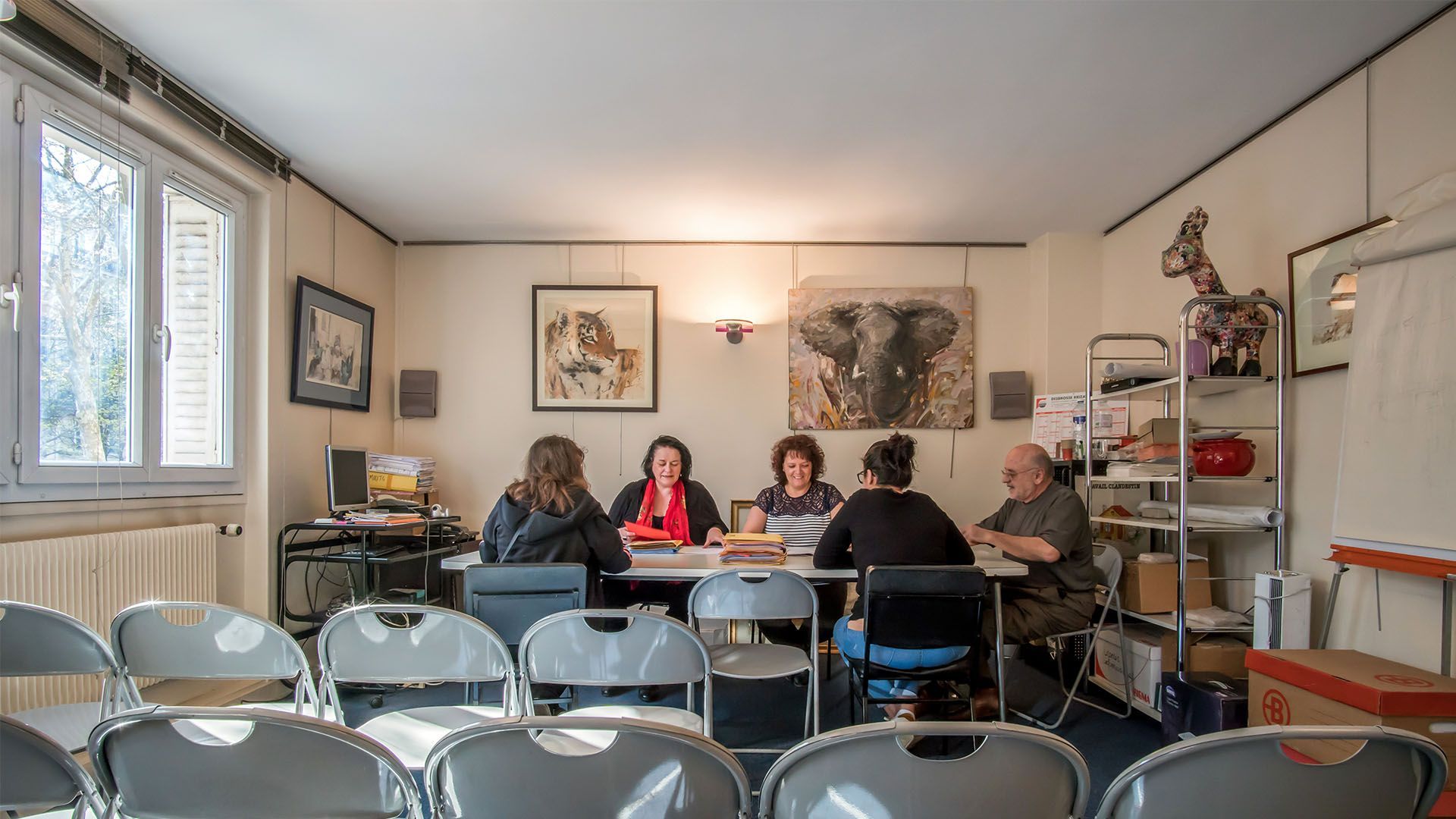 Des personnes sont assises à une table dans une pièce dont les murs sont décorés d'œuvres d'art. Des chaises pliantes grises font face à la table.