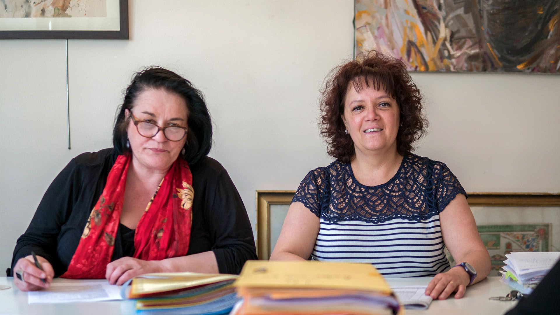 Deux femmes assises à une table, l'une écrit, l'autre sourit. Des papiers et des œuvres d'art sont visibles en arrière-plan.