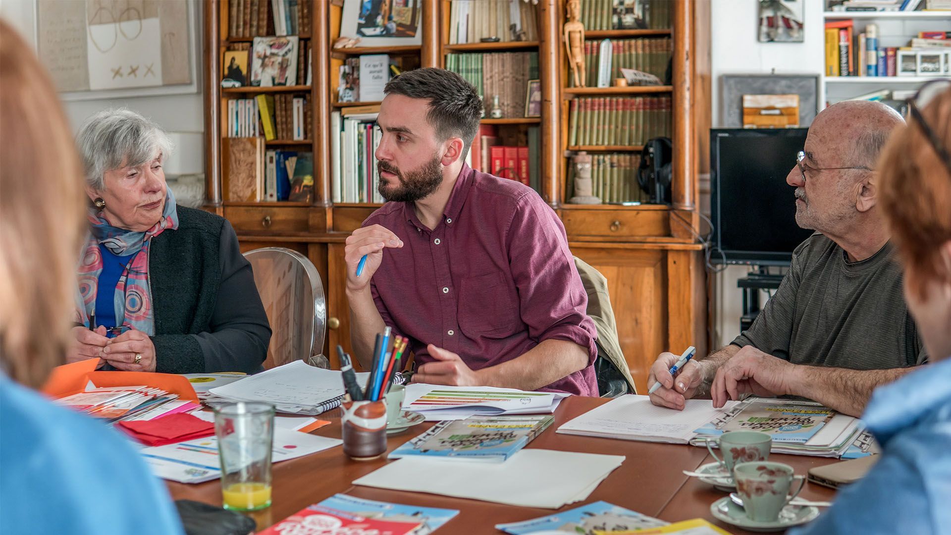Des personnes assises à une table discutent. Un homme en chemise bordeaux fait un geste. On aperçoit des étagères et un bureau avec des papiers.