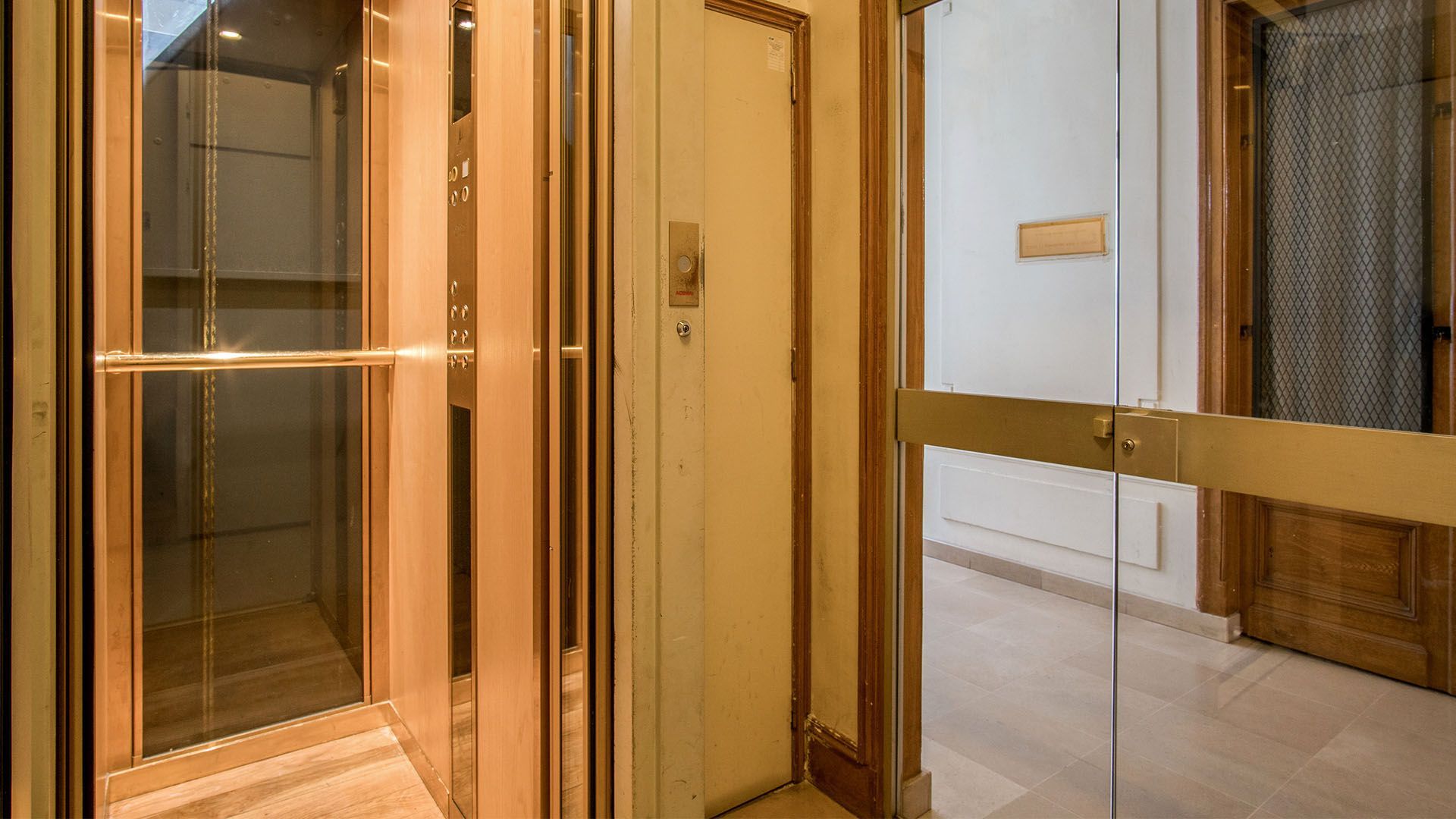 Un ascenseur aux finitions dorées et à porte vitrée jouxte une porte fermée et un couloir.