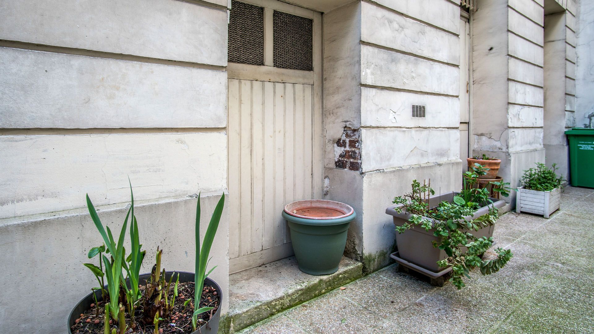 Porte d'entrée extérieure avec murs blancs patinés, plantes en pot et poubelle verte.