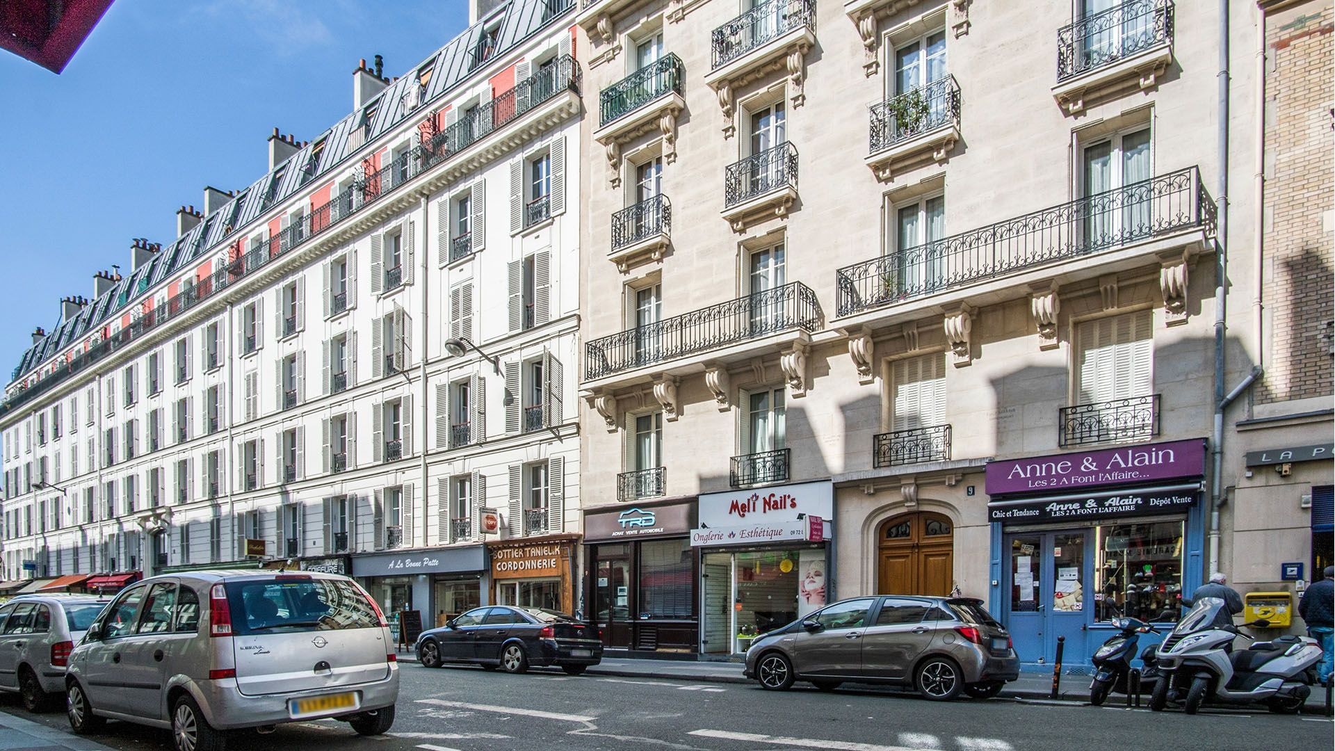 Vue de rue d'immeubles parisiens, voitures garées dans la rue, magasins en contrebas, balcons aux étages supérieurs.