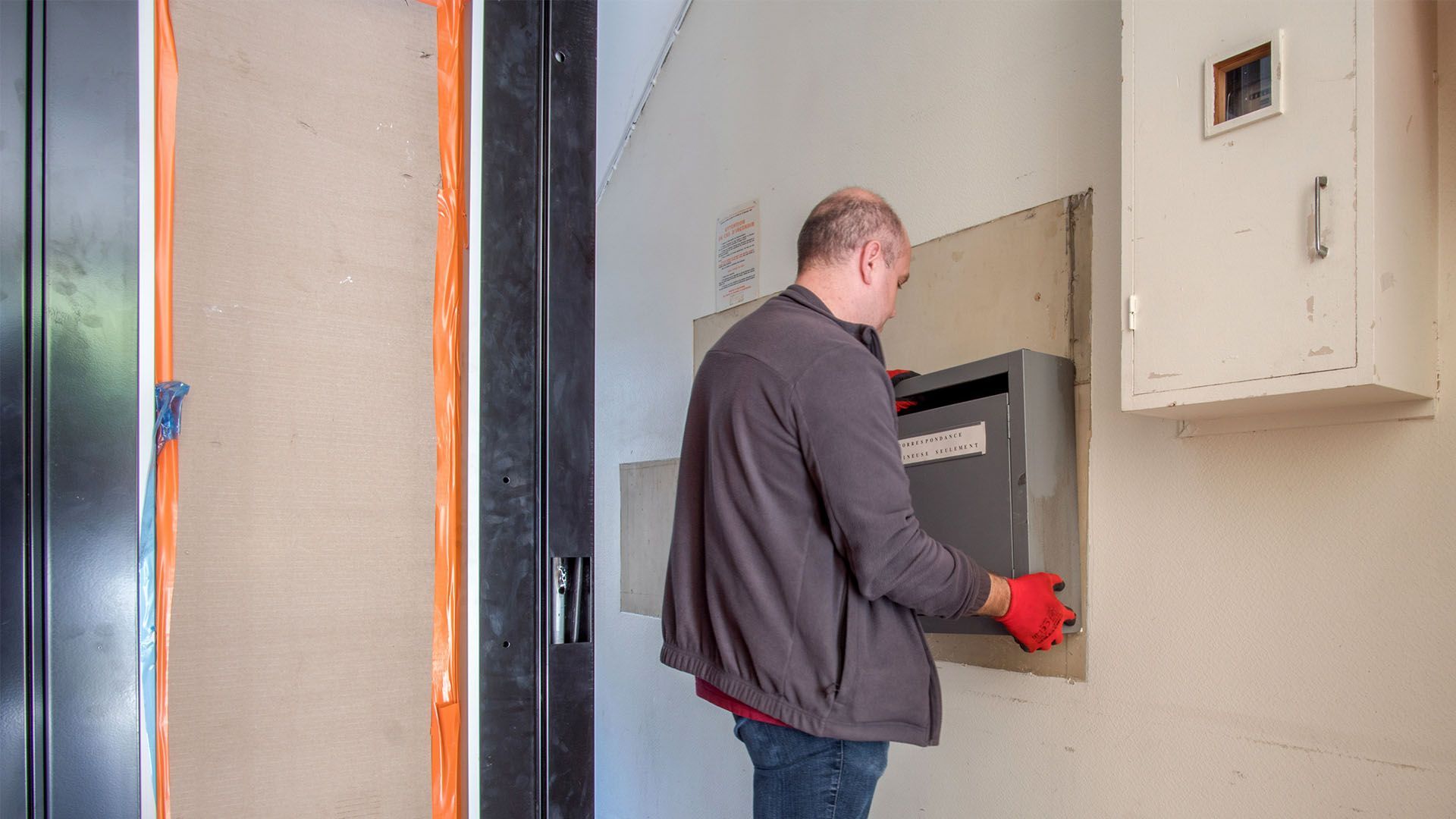 Un homme en veste grise et jean travaille sur un tableau électrique mural. On aperçoit des gants rouges.