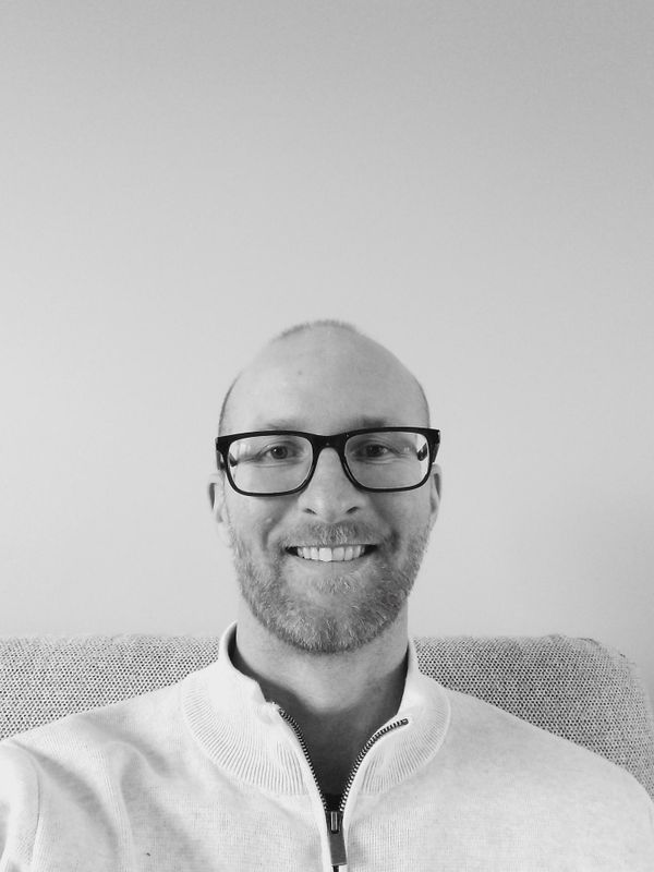 Un homme chauve souriant, portant des lunettes et un pull blanc à fermeture éclair, est assis contre un mur clair.