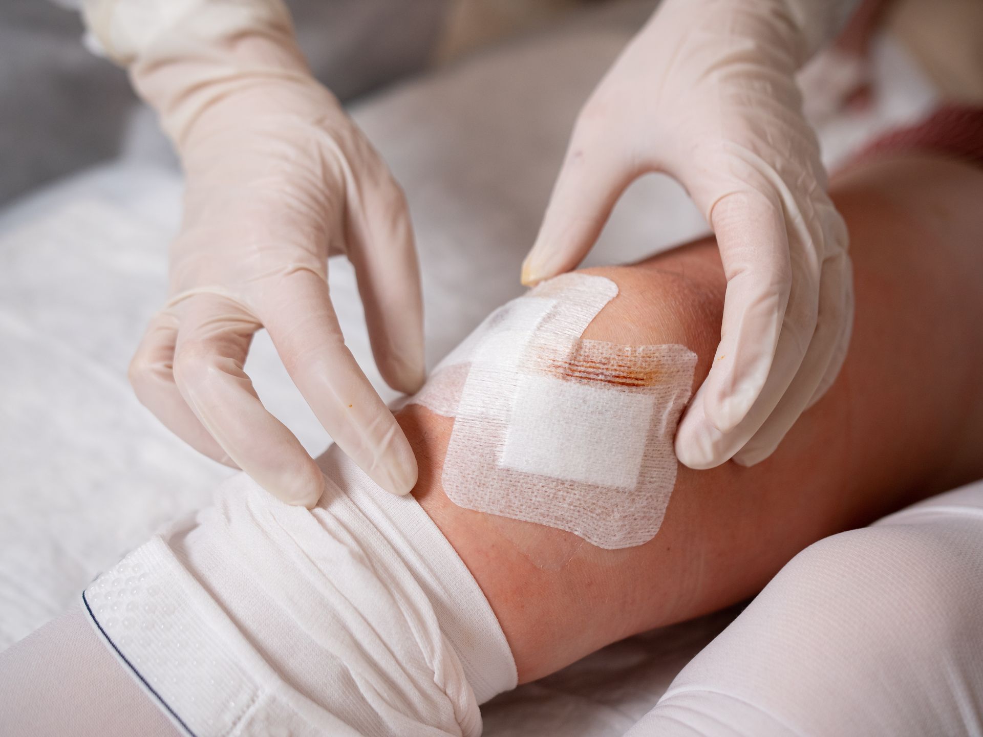 Des mains gantées appliquent un bandage de gaze sur le genou d'un patient allongé sur un lit.