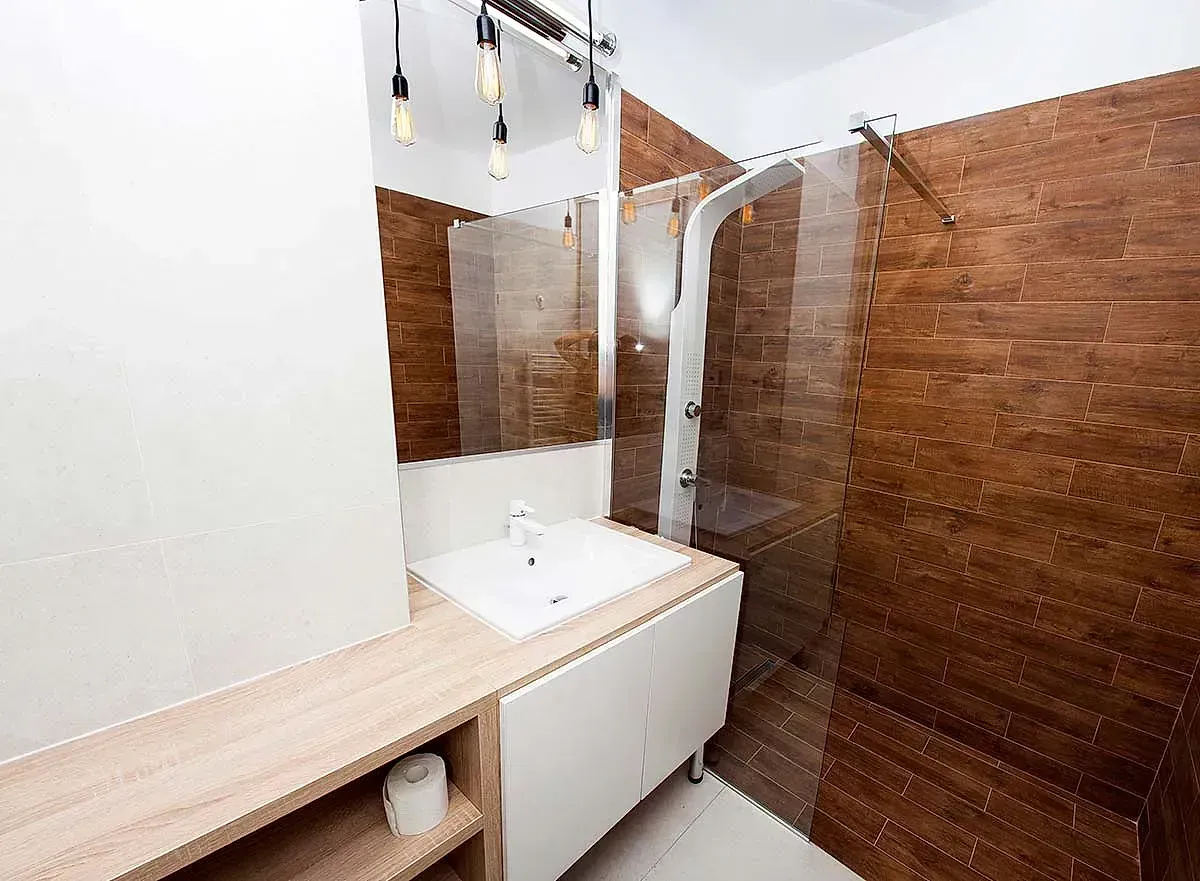 Baño con azulejos de apariencia madera, lavabo y tocador blancos, ducha y artefactos de iluminación modernos.