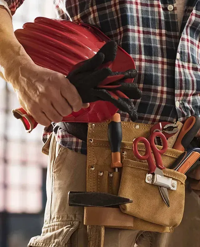 Una persona con un cinturón de herramientas sosteniendo un casco rojo y guantes negros.