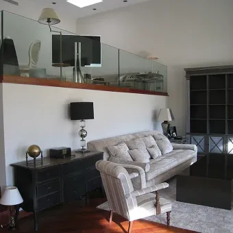 Sala con sofá, sillón, mueble y TV en entrepiso con barandales de vidrio.
