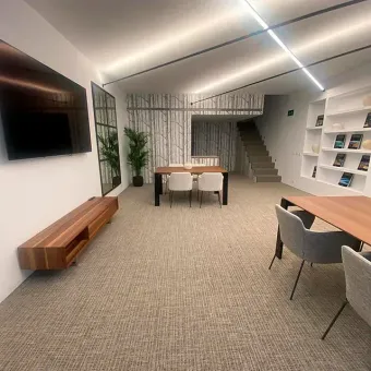 Oficina moderna con mesas, sillas, televisor y árbol decorativo. Colores neutros, iluminación empotrada y escaleras.