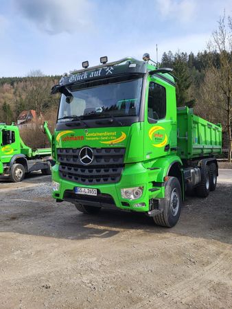 Grüner Muldenkipper auf Schotter geparkt, im Hintergrund ein zweiter LKW. Mercedes-Logo auf der Vorderseite.