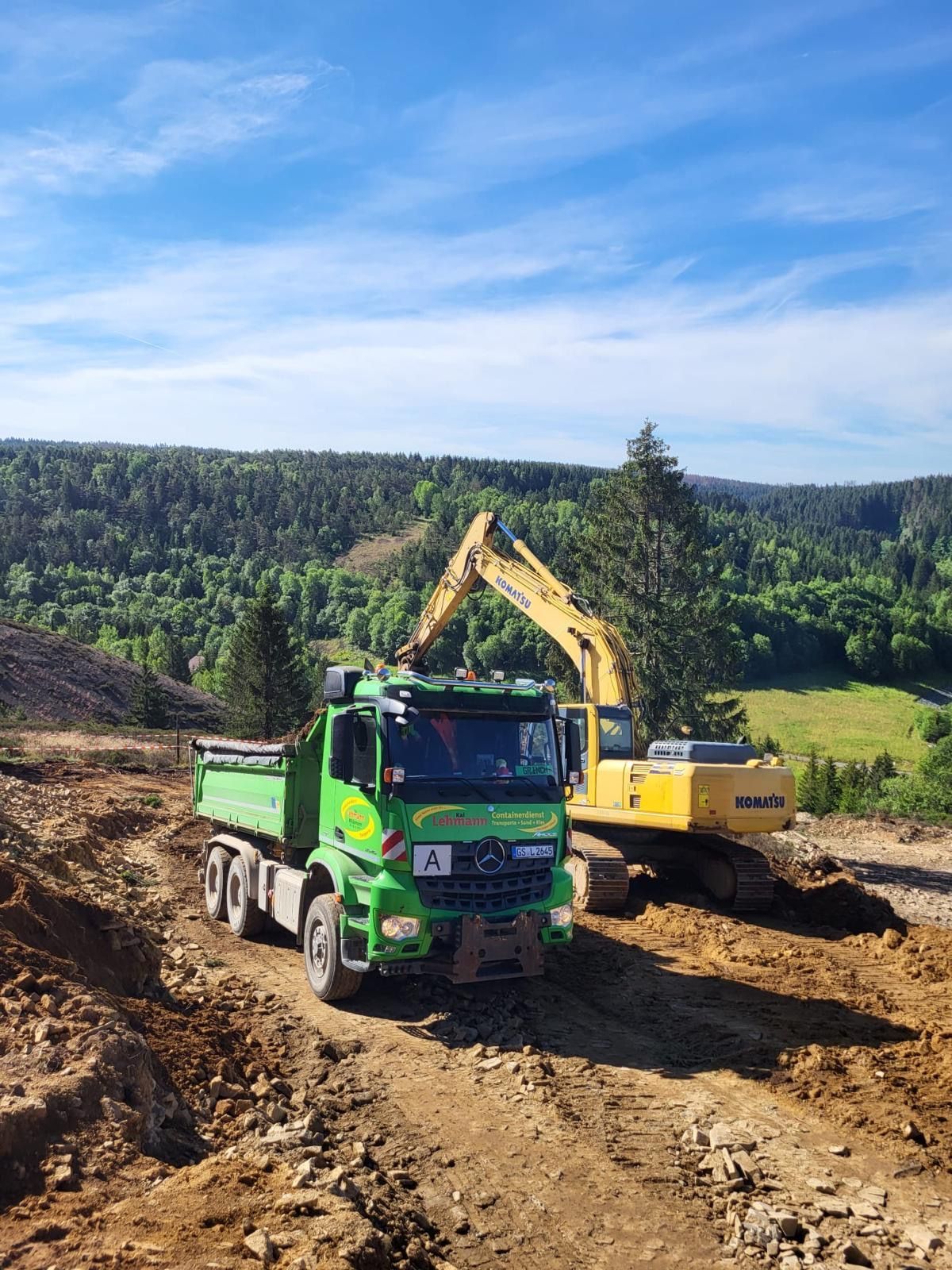 Grüner Muldenkipper parkt neben einem gelben Bagger auf einer unbefestigten Straße, Wald im Hintergrund, blauer Himmel.