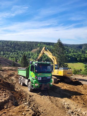 Grüner Muldenkipper parkt neben einem gelben Bagger auf einer unbefestigten Straße, Wald im Hintergrund, blauer Himmel.