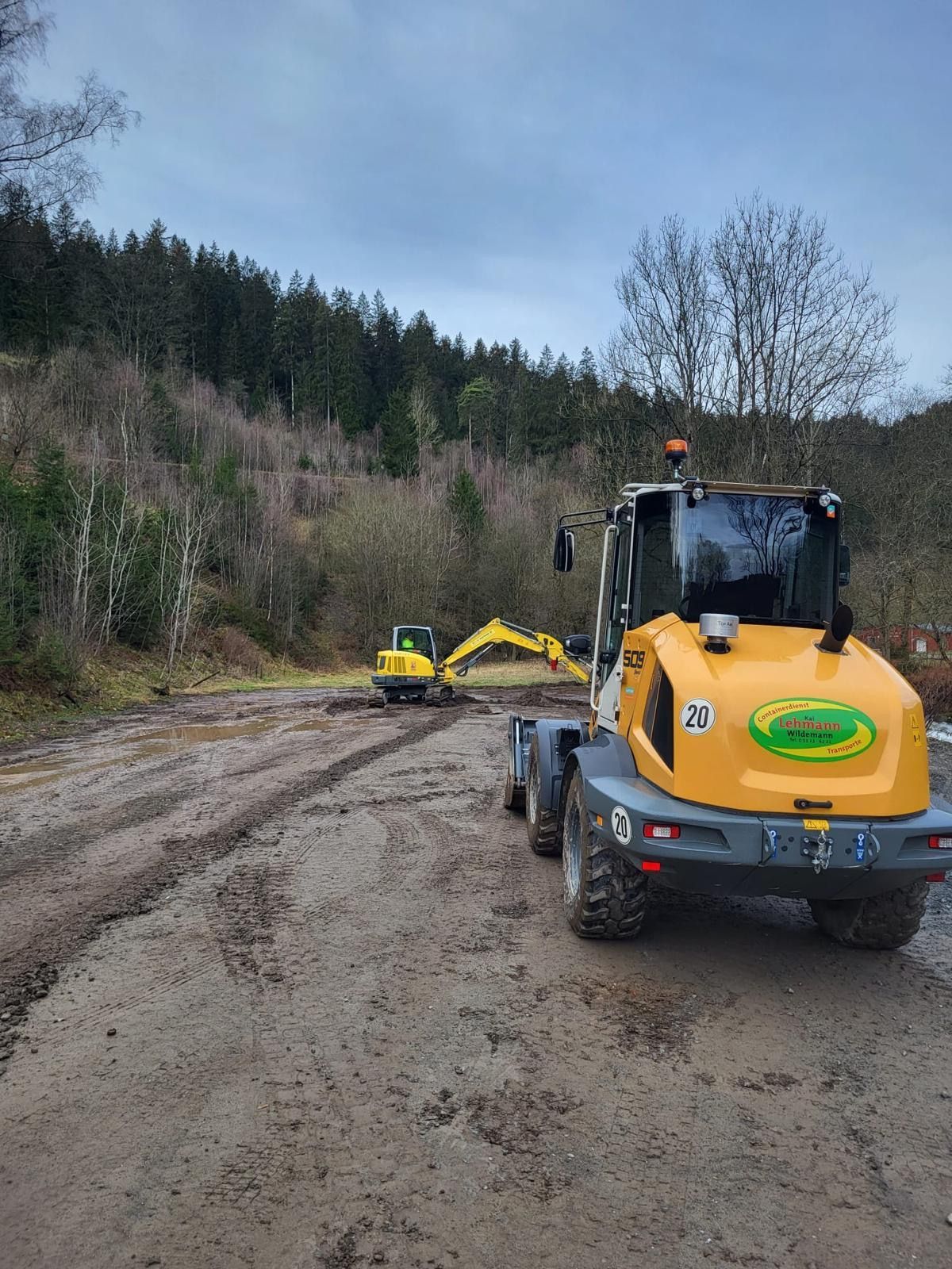Gelbes Baufahrzeug auf schlammigem Boden mit Baggern im Hintergrund, Waldkulisse.