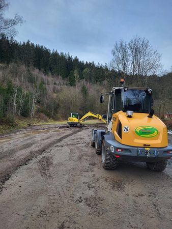 Gelbes Baufahrzeug auf schlammigem Boden mit Baggern im Hintergrund, Waldkulisse.