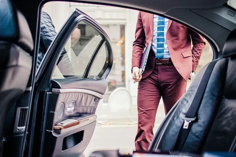 Hombre con traje saliendo de un automóvil; puerta abierta, entorno empresarial, traje marrón, sosteniendo una computadora portátil.
