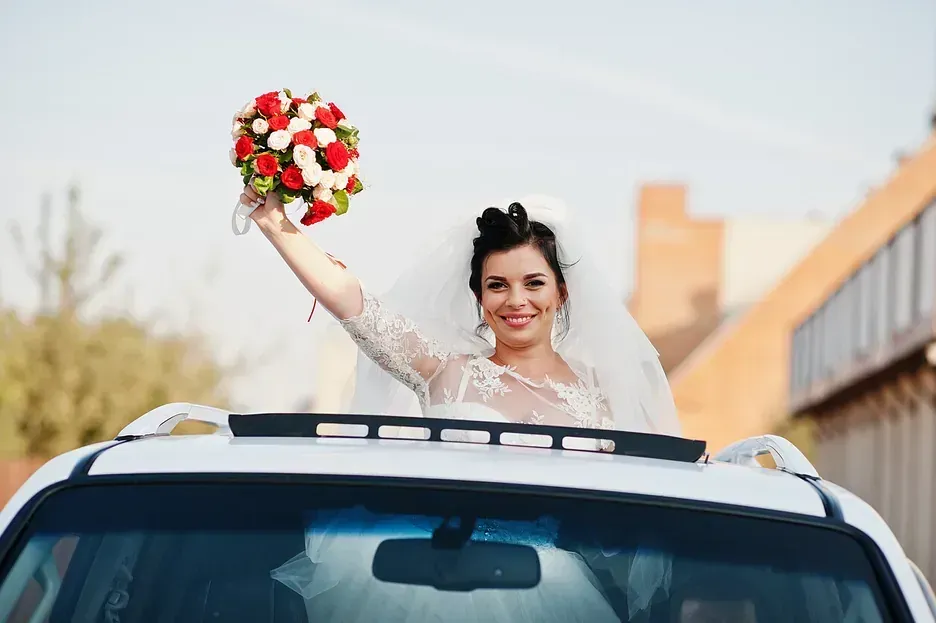 Una novia saluda desde el techo corredizo de un coche, con un ramo en la mano. Sonriendo, con velo y vestido de encaje, al aire libre.