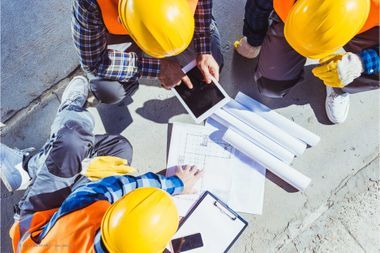 Drei Bauarbeiter mit gelben Helmen sitzen über dem Bauplan