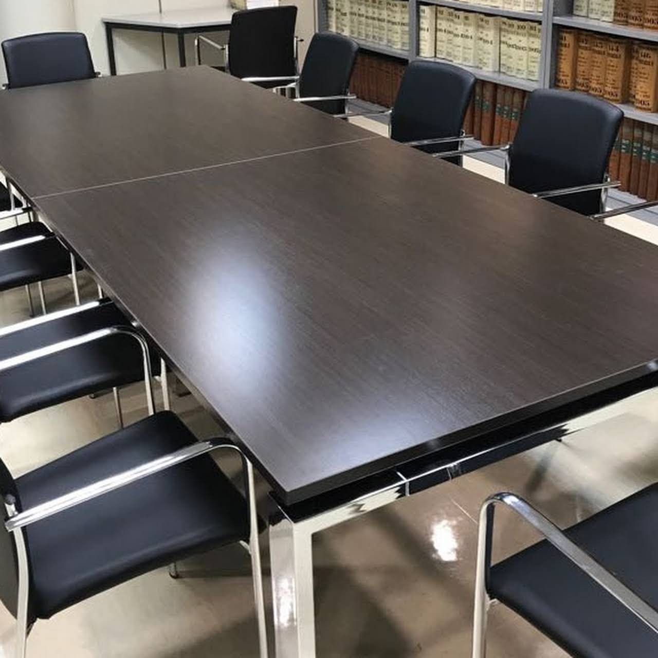 Sala de conferencias con una mesa larga de madera oscura y sillas negras; estanterías al fondo.