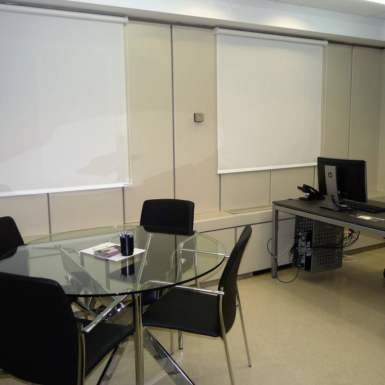 Interior de oficina con una mesa de vidrio, sillas negras, escritorio de computadora y cortinas blancas.