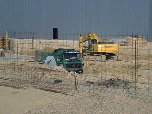 Un sitio de construcción con un camión verde y una excavadora amarilla detrás de una valla.