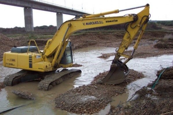 Una excavadora amarilla está parada en el barro cerca de un puente.