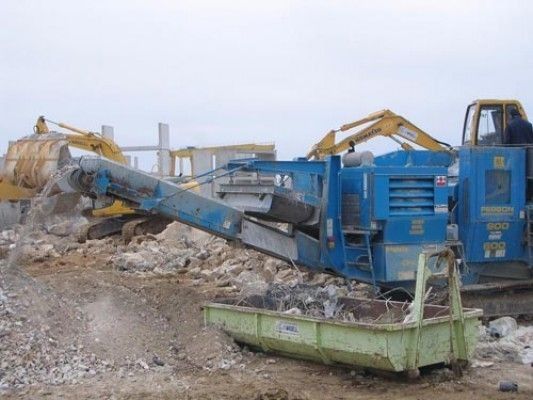 Un sitio de construcción con una máquina azul y un contenedor de basura verde.