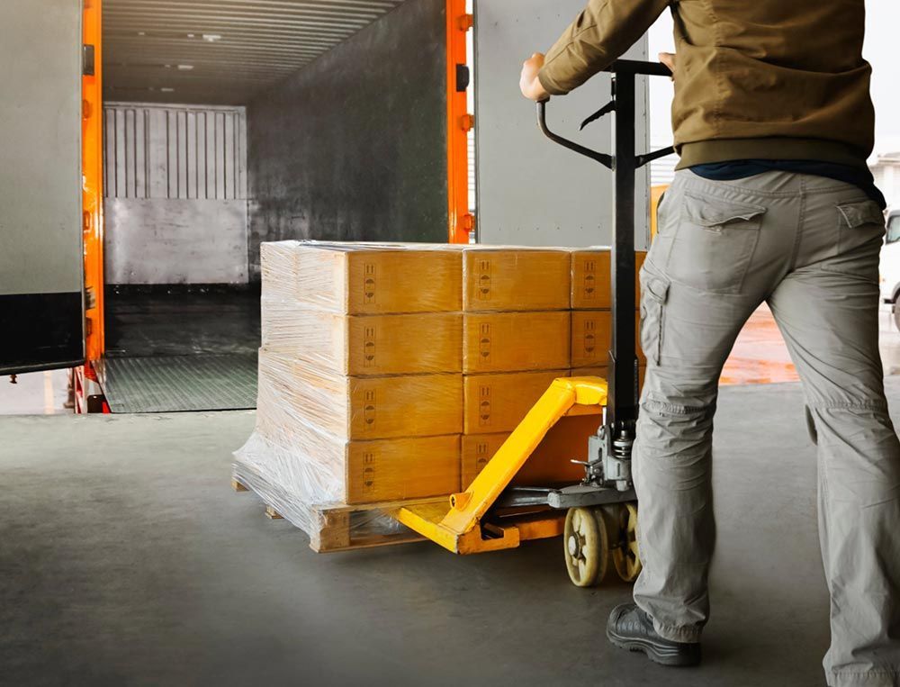 Un trabajador empuja una transpaleta con cajas apiladas hacia una zona de carga de camiones.
