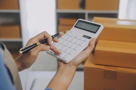 Manos usando una calculadora junto a cajas de cartón apiladas en un espacio de trabajo de envío.