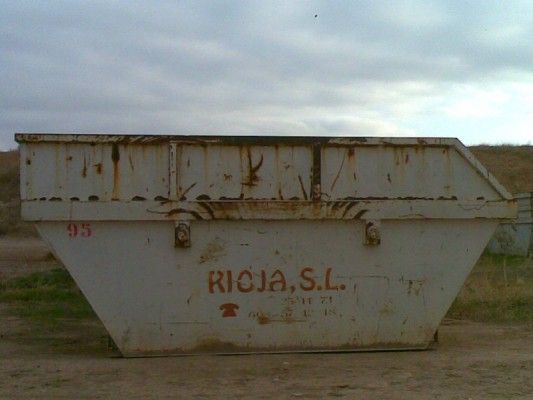 Un contenedor industrial blanco oxidado con la etiqueta “RICJA, SL” en un campo al aire libre bajo un cielo nublado.