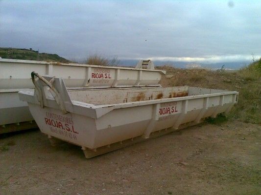 Un gran contenedor de obra blanco lleno de escombros en un terreno baldío bajo un cielo nublado.