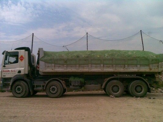 Camión volquete cargado de material verde en un camino de tierra bajo un cielo nublado.