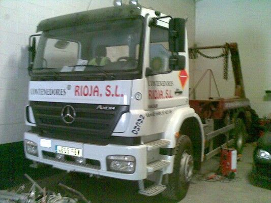 Camión Mercedes blanco estacionado en el interior con el logotipo de la empresa y un brazo de grúa en la parte trasera.