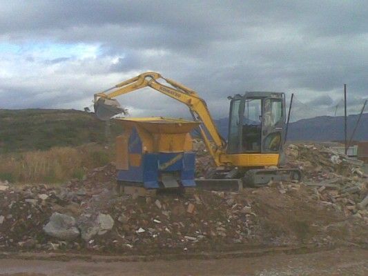 Una excavadora amarilla carga escombros en un contenedor azul en una obra de construcción bajo un cielo nublado.