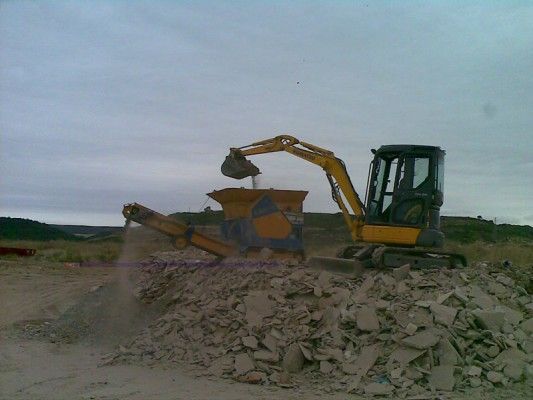 Excavadora cargando escombros en una trituradora en una obra de construcción bajo un cielo nublado.