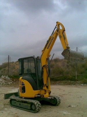 Miniexcavadora amarilla sobre orugas levantando una pala en un terreno baldío bajo un cielo nublado.