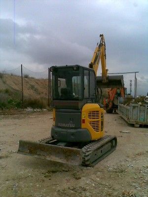 Una pequeña miniexcavadora amarilla en un terreno de grava, con una excavadora más grande y una valla al fondo.