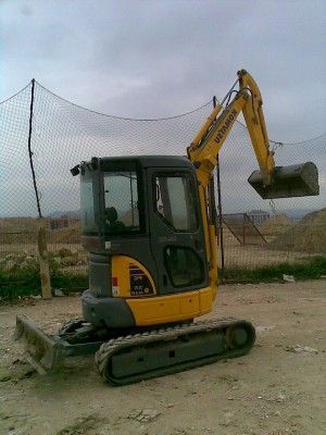 Miniexcavadora amarilla estacionada en un terreno de tierra con su pala elevada cerca de una valla de red.