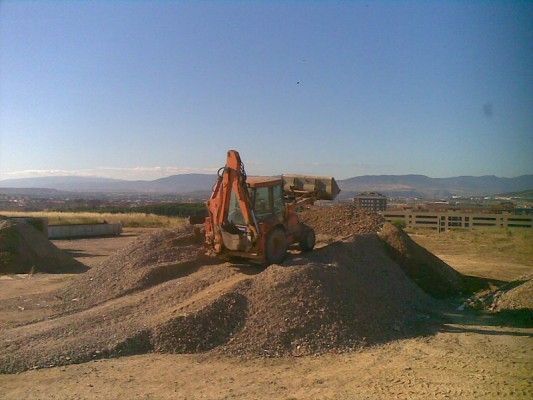 Excavadora naranja sobre un montículo de tierra en una obra de construcción bajo un cielo azul despejado.