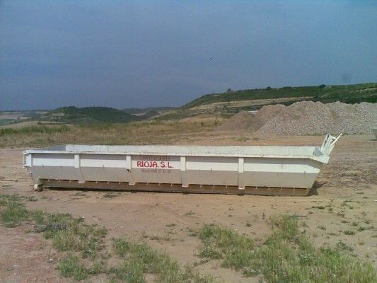 Contenedor de obra blanco sobre terreno arenoso junto a montículos de tierra bajo un cielo despejado.