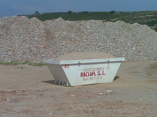 Contenedor de obra blanco con la etiqueta “RIJOA, SL” en un terreno de grava con una gran pila de escombros detrás.