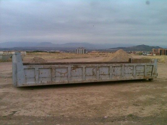 Un gran contenedor de basura gris en un terreno baldío con montañas al fondo.