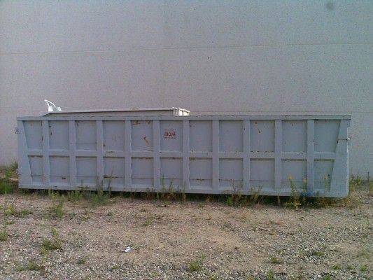 Un gran contenedor de basura metálico gris sobre grava junto a una pared lisa.