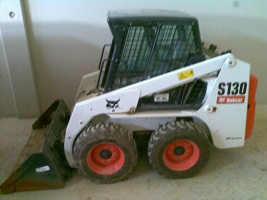 Minicargadora Bobcat blanca con ruedas rojas y pala frontal en el interior, cerca de una pared.