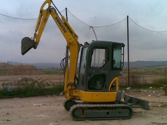 Excavadora amarilla en un terreno baldío junto a una cerca de alambre bajo un cielo nublado.