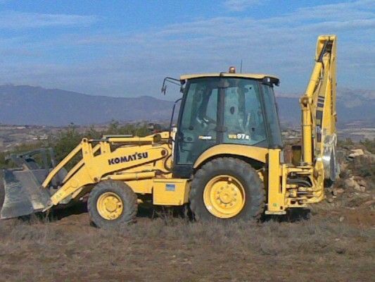 Retroexcavadora amarilla estacionada en un campo de tierra con montañas al fondo.