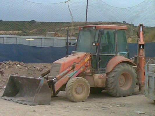 Retroexcavadora naranja en una obra de construcción junto a un montón de tierra y una valla.